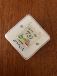 An Aranet4 carbon dioxide monitor: A small white square device about the size of a human palm, with an e-ink display showing the PPM of carbon dioxide, the temperature, and the relative humidity.